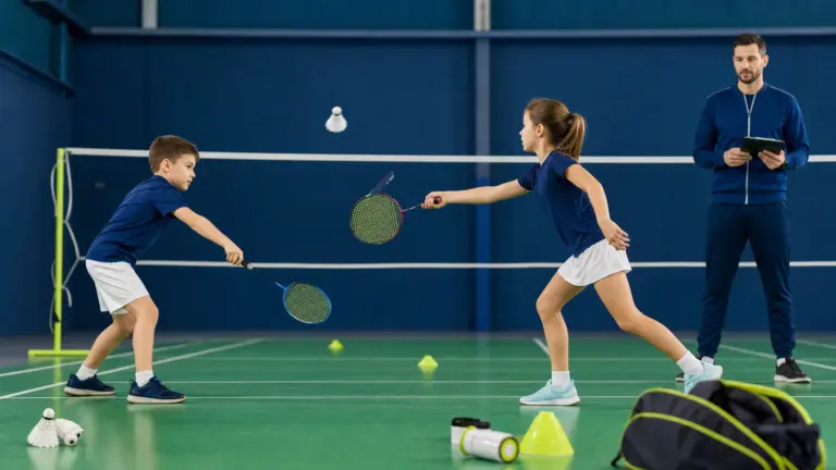 Badminton filesi önünde raketlerini tüytopa uzatan iki çocuk ve tablet tutarak izleyen bir yetişkin